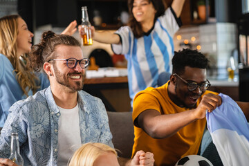 Excited group of people watching football, soccer sports match at home. Multi-ethnic group of emotional friend fans cheering for the favorite national team, drinking beer. Multiethnic friends hanging