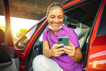 A smiling young nurse dressed in her scrubs uniform sitting in her car holding her mobile phone,...