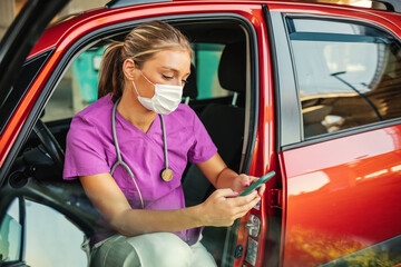 A photo of a young female doctor sitting in her car wearing a protective mask and looking at her...