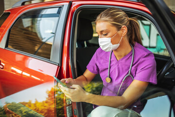 A photo of a young female doctor sitting in her car wearing a protective mask and looking at her...
