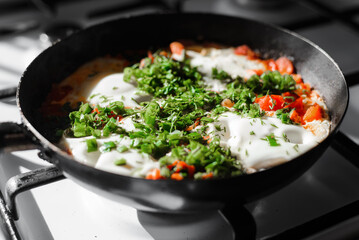 Shakshuka is dish of eggs fried in sauce of tomatoes, hot peppers, onions and spices in frying pan on gas stove in kitchen. Close-up, selective focus