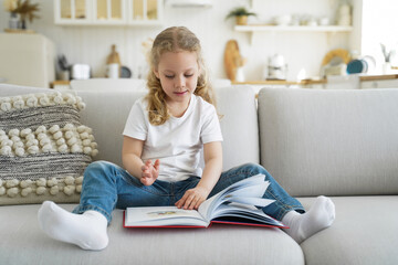 Little girl is sitting on couch and reading fairy tale book. Preschool child is learning to read.