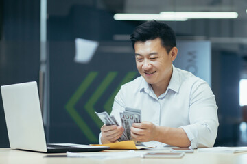Successful asian investor counting dollars money cash sitting in office, happy man smiling and happy