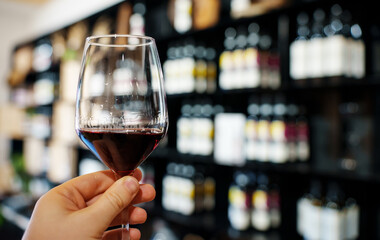 Man's hand holds glass with red wine on the background of bottles.