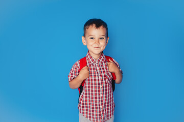 A cheerful smiling little brunette boy with a backpack is having fun on the background of a blue wall. I look into the camera. The concept of the school. back to school