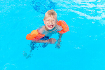 Fototapeta na wymiar happy baby boy swims or learns to swim in the blue water pool in armbands, the concept of summer holidays and school holidays