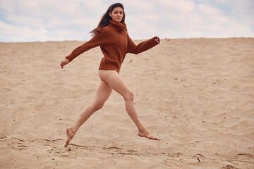 Beautiful woman jump up with windy hair on sandy beach. Stylish young sexy female in knitted sweater and bare legs relaxing on coast.