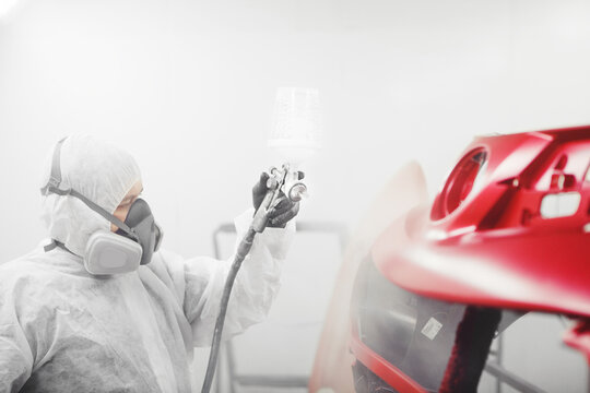Man in protective mask and clothes sprays varnish to car bumper with a spray gun in a paint booth.