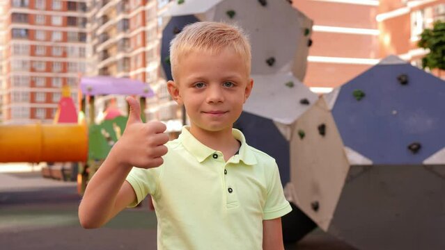 A little smiling eight-year-old boy stands on the playground, looks at the camera and shows a thumbs up.