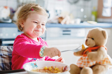 Adorable baby girl eating from fork vegetables and pasta. food, child, feeding and development concept. Cute toddler, daughter with spoon sitting in highchair and learning to eat by itself.