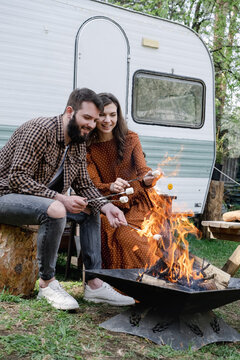 Happy young caucasian couple is traveling in travel van. Romantic atmosphere of relaxation. Road trip around country for weekend. Man and woman roasting marshmallows on campfire. Millennial generation