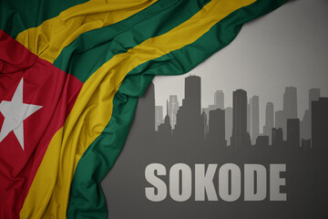 abstract silhouette of the city with text sokode near waving colorful national flag of togo on a gray background.