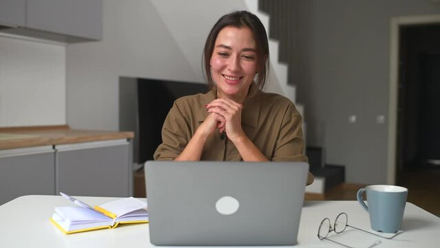 Smiling asian businesswoman taking notes sitting at the desk in modern home office space. Young multiracial female student overwrites from the laptop screen into notebook