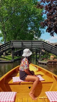 Asian woman with hat in a wooden boat at the canals of Giethoorn Netherlands. Giethoorn, Netherlands: View of famous Giethoorn village with canals and rustic thatched roof houses