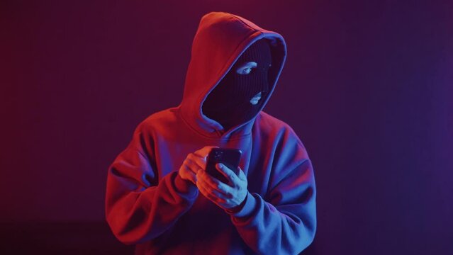 man in a balaclava in a dark room talking on a smartphone