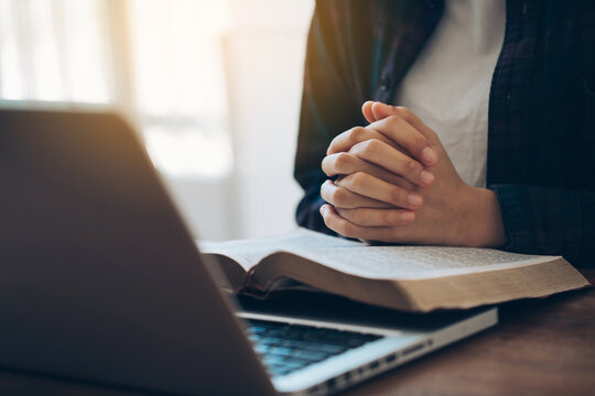 Women's hands are folded in prayer on a Holy Bible over a laptop in church concept for faith, religion, love, and forgiveness. Holy Bible study reading together in Sunday school.Online church