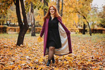 Outdoor autumn portrait of happy smiling plus size red hair woman in coat walking in fall park.
