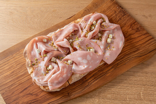 A piece of a traditional pizza of the Roman tradition, known as Pinsa Romana, with Mortadella of Bologna, Gorgonzola and Pistacchio grains, vision from above on wooden table and cutting board 