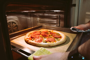 A man's hands pull out a homemade pepperoni pizza from the oven