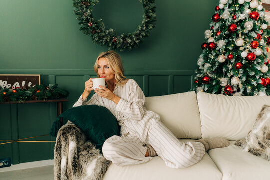 Gorgeous caucasian blond woman sitting on sofa on Christmas morning drinking coffee enjoying