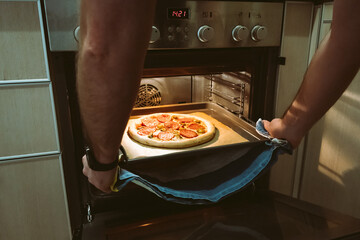 A man bakes pizza at home using an oven. Men's hands pull a pizza baking sheet out of the oven.
