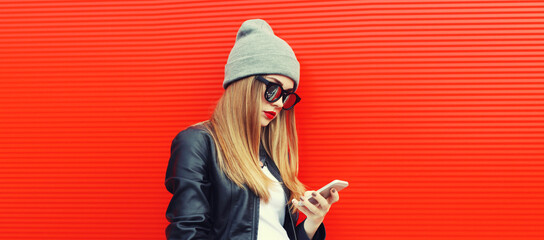 Portrait of stylish woman with smartphone wearing gray hat, leather biker rock jacket on red background, blank copy space for advertising text
