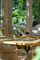Two glasses of vintage red wine stand on wooden table in glare of sun. Cozy interior of terrace of summer cafe. Soft cushions on chair for comfort. Green plants outside windows. Romantic relax concept
