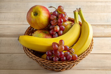 Ripe fruits in wicker basket on wooden background. Bananas, apples and grapes. Harvest concept.