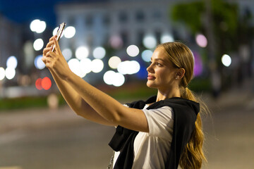 Young woman using mobile smartphone on background bokeh light in night city, woman taking selfie photo, online wi-fi internet