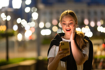 Amazed young woman checking smart phone content sitting in the street in the night