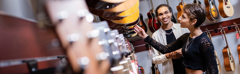 Vendeur afro-américain regardant une femme choisissant une guitare acoustique dans un magasin de musique, bannière.