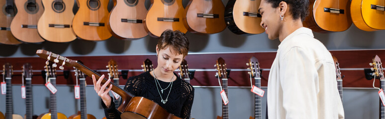 Woman holding acoustic guitar near smiling african american seller in music store, banner.