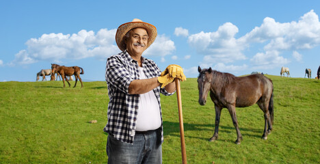 Farmer with a straw hat leaning on a wooden stick on a field with horses