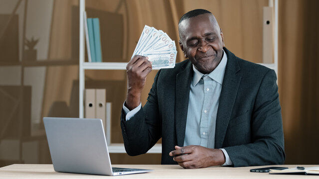 Old Mature African Businessman 60s Worker Banker Banking Agent Senior Man Sitting In Office Rejoices In Financial Win Holds Fan Of Money Sniffing Dollars Satisfied With Salary Loan Wealth Success