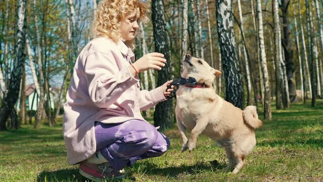 Attractive pretty caucasian woman wearing a pink coat crouching on the ground and training her small golden dog in a park.