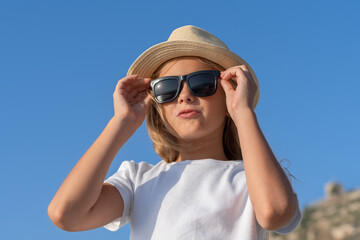 A young girl in a hat and sunglasses adjusts her glasses with two hands.