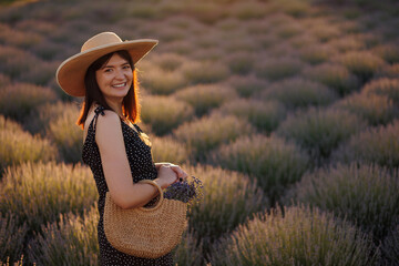 Cheerful woman in lavender field with flowers in bag during sunset