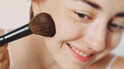 A young white beautiful girl with a makeup brush puts makeup on her face. Facial powder.