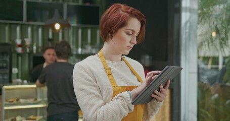 Fototapeta premium Young employee woman wearing apron standing inside coffee shop holding tablet device checking online orders. Red hair girl working at cafe