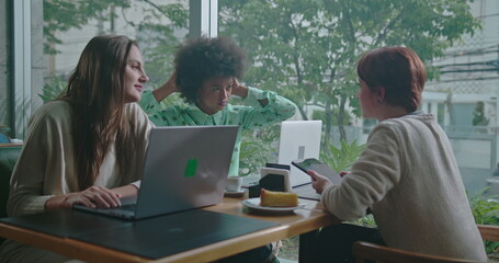 Fototapeta premium Friends sitting at table inside coffee shop talking. Three female diverse colleagues in conversation sharing gossip and chatting