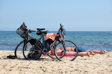 Fototapeta premium A bike trip to the beach on a summer day - two bikes are standing on the seashore. Two people are lying on the sand behind the bicycles and sunbathing.