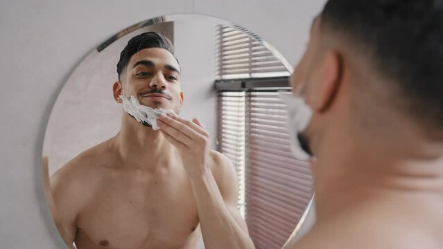 Male reflection in mirror in bath Arabian handsome Indian bearded man Hispanic guy applies white foam shaving gel male cream preshave cosmetics to beard preparing to shaves dermatology facial care