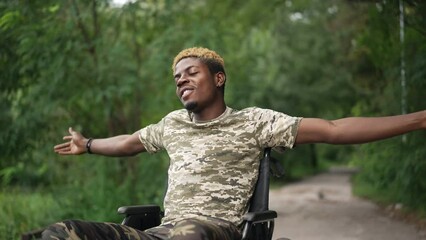 Happy man in wheelchair stretching hands with closed eyes enjoying tranquility in forest. Medium shot portrait of smiling African American guy resting outdoors in green park. Disability individuality