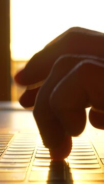 TRZIN, SLOVENIA, 20.5.2022: SLOW MOTION, CLOSE UP: Hands typing on laptop keyboard in early morning sunlight. Sunrise rays shining over fingers of a typing person and laptop keyboard in home office