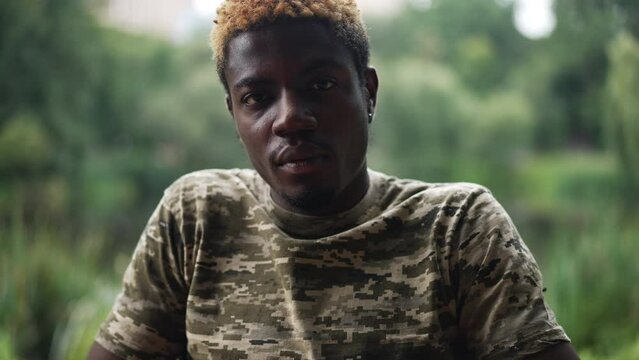 Portrait of young African American man in khaki T-shirt looking at camera with serious facial expression as live camera zoom in. Confident courageous soldier in wheelchair posing in park outdoors