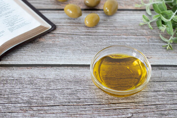 Pure olive oil in a glass container, olives, green branch, and an open Holy Bible Book on a wooden...