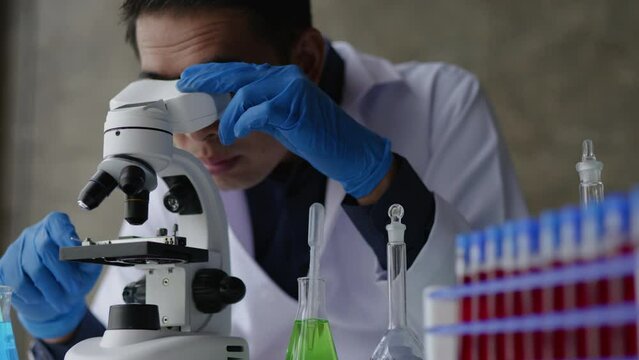 Male scientist using microscope for observation on simple test in laboratory table that full of test tubes and beakers.