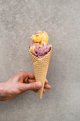Man's hand holding melting vanilla ice cream with pink and yellow scoops in waffle cone on gray...