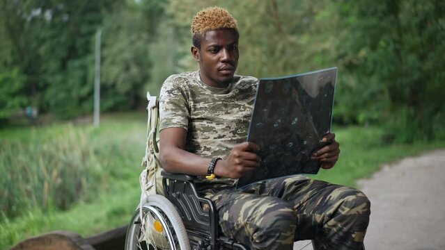 Sad thoughtful young African American soldier in wheelchair examining X-ray sitting in summer park. Portrait of devastated hopeless handsome man thinking procrastinating outdoors in slow motion