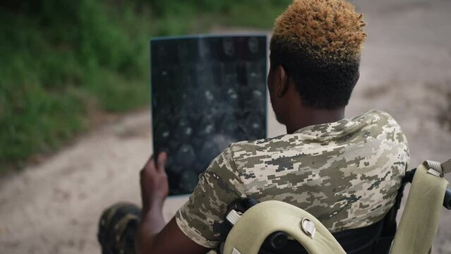 Shooting over shoulder of young man in wheelchair with X-ray image thinking sitting outdoors. African American sick soldier analyzing medical result in summer park forest. Slow motion
